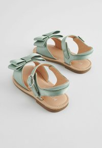 Sandali verde menta con una texture satinata, caratterizzati da un grande fiocco decorativo, cinturino alla caviglia regolabile e suola beige.