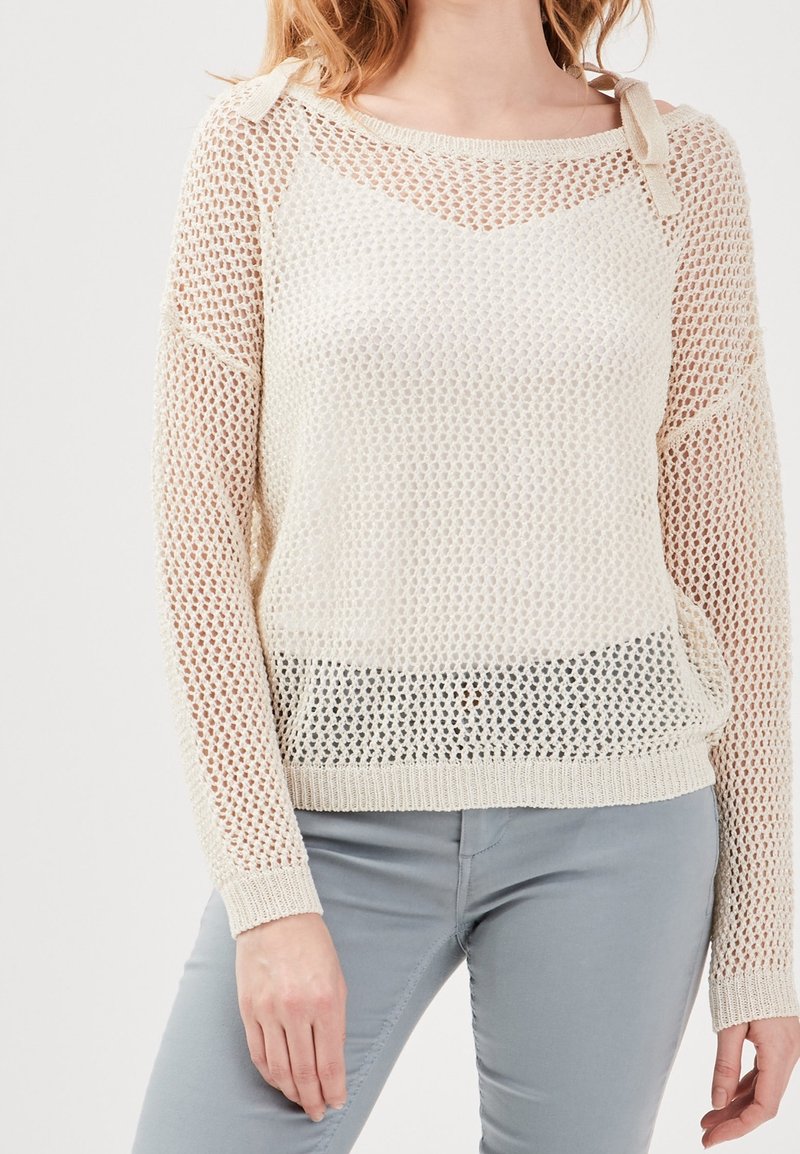 Pull en tricot beige clair à maille lâche, présentant un motif texturé et des nouettes en ruban aux épaules. Manches longues et ourlet côtelé.