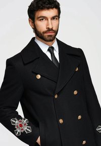 Schwarzer Woll-Doppelreiher-Mantel mit goldenen Knöpfen und dekorativer Stickerei an den Ärmeln mit roten und weißen Verzierungen.