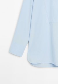 Camicia azzurra a maniche lunghe con maniche rialzate e orlo curvo su sfondo bianco.