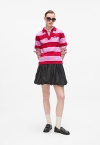 Polo rayé rose et rouge avec un col en V, associé à une jupe plissée noire. Le mannequin porte des lunettes de soleil et des mocassins noirs avec des chaussettes.