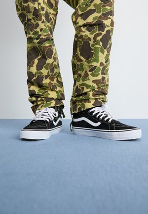 Svart sneakers med hvite detaljer, med en teksturert overdel og kontraststing. Kombinert med grønne camouflagemønstrede bukser.