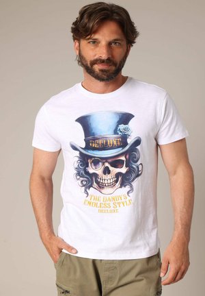 T-shirt en coton blanc avec un imprimé graphique d'une skull portant un haut-de-forme, des accents dorés, et le texte "LE STYLE SANS FIN DU DANDY."