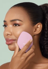 Dispositif de nettoyage facial rose tenu contre la joue, présentant un design lisse et arrondi avec des boutons et un logo sur la surface.