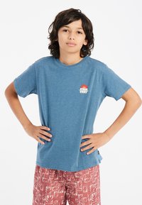 Mėlynas trumpomis rankovėmis T-shirt su apvaliu iškirpimu ir mažu grafiniu logotipu. Sujungta su raštuotomis raudonomis šortais, turinčiomis lapų dizainą.