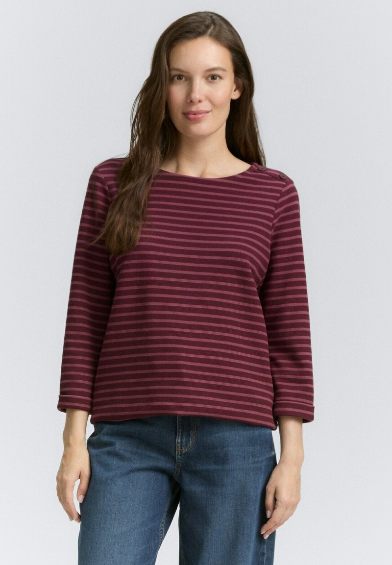 Langes Oberteil mit burgunderroten und rosa Streifen, rundem Halsausschnitt und Knopfdetails an den Schultern, kombiniert mit blauen Denim-Hosen.