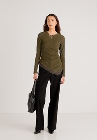 Ilgos rankovės alyvuogių žalios spalvos palaidinė su tekstūruota medžiaga ir nėriniuota apačia, derinama su juodomis platėjusiomis kelnėmis ir blizgia juoda rankine.