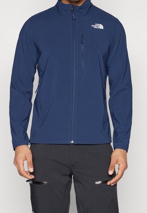 Mann trägt eine marineblaue The North Face Jacke mit Reißverschluss und Reißverschlusstaschen an den Seiten und auf der Brust, kombiniert mit schwarzen Hosen mit Reißverschlusstaschen am Oberschenkel.