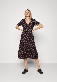Vestido floral preto com mangas curtas, decote em V, comprimento até a metade da panturrilha, com flores vermelhas e verdes, e tecido leve. Combinado com sapatos pretos tipo slip-on.