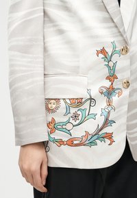 Blazer beige clair avec rayures subtiles, boutons dorés et broderie florale colorée orange, bleu sarcelle et bleu près de la poche et de l'ourlet.