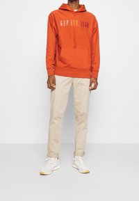 Felpa arancione con logo "GAP EST. 1969" in colori gradient, tasca frontale, abbinata a pantaloni beige e sneakers bianche con accenti grigi.