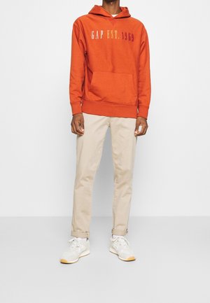 Sudadera naranja con el logo "GAP EST. 1969" en colores degradados, con bolsillo frontal, combinada con pantalones beige y zapatillas blancas con acentos grises.