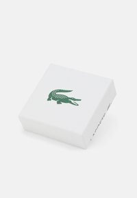 Balts dāvanu kaste ar teksturētu virsmu, ar zaļu krokodila logotipu izcili redzamu uz vāka un uz sāniem izdrukātu "LACOSTE".