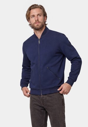 Man wearing a navy blue zip-up jacket with side pockets and dark gray pants, looking off to the side against a plain background.
