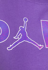 Fialové bavlněné tričko s bílou siluetou skákající postavy s basketbalem, zvýrazněné růžovým třpytem. Velká písmena.