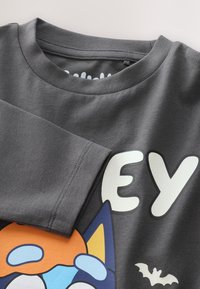 Graues Baumwoll-T-Shirt mit einem bunten Cartoon-Design, das eine orangefarbene Figur, große weiße Buchstaben und eine kleine Fledermausgrafik zeigt.