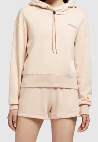 Perzikkleurige hoodie met trekkoorden en een kangoeroezak, gecombineerd met bijpassende shorts. Beide items hebben een zachte, gestructureerde afwerking en logo-accenten.