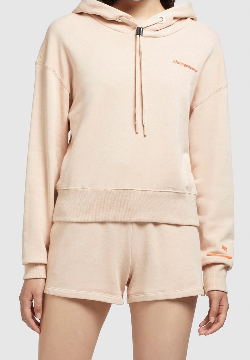 Perzikkleurige hoodie met trekkoorden en een kangoeroezak, gecombineerd met bijpassende shorts. Beide items hebben een zachte, gestructureerde afwerking en logo-accenten.
