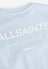 Gaiši zils kokvilnas T-krekls ar mīkstu apaļu kakla izgriezumu un lieliem baltiem uzrakstiem "ALLSAINTS" un "LONDONA" dažādās valodās.