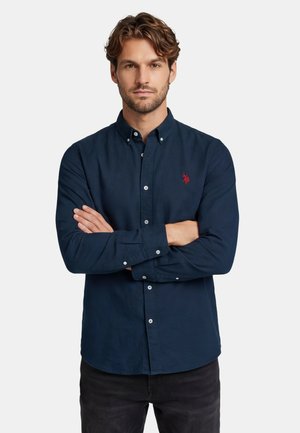 Man with short wavy hair wearing a navy blue buttoned shirt with a red embroidered logo, standing with arms crossed against a white background.