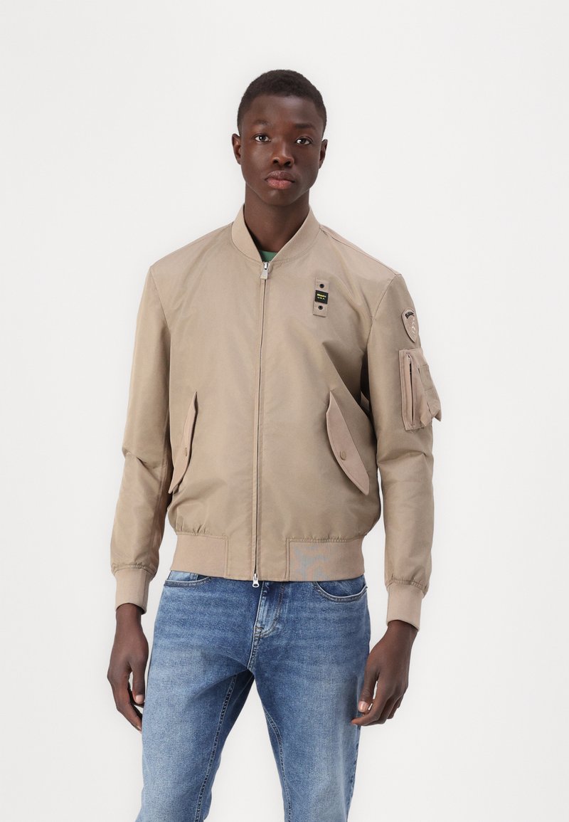Beige bomberjakke med glidelås, side lommer og lomme på ermet. Har ribbede mansjetter og en glatt, lett tekstur.