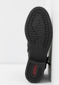 Semelle de chaussure noire en caoutchouc texturé, présentant un motif géométrique et un logo rouge. La vue latérale révèle une construction robuste et une forme.