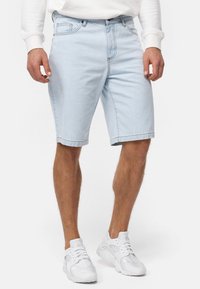 Shorts en denim bleu clair avec une coupe droite, comportant cinq poches, des passants de ceinture et un ourlet surpiqué. Portés avec des chaussures de sport blanches.