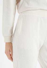 Białe spodnie dresowe z polaru z wytłaczanym wzorem gwiazdy, wyposażone w prążkowany pas i mankiety. Materiał ma miękkie, teksturowane wykończenie.