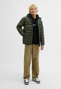 Grön vadderad jacka med huva, svart hoodie under, khakifärgade byxor och svart-vita sneakers. Har en kviltad design och fickor.