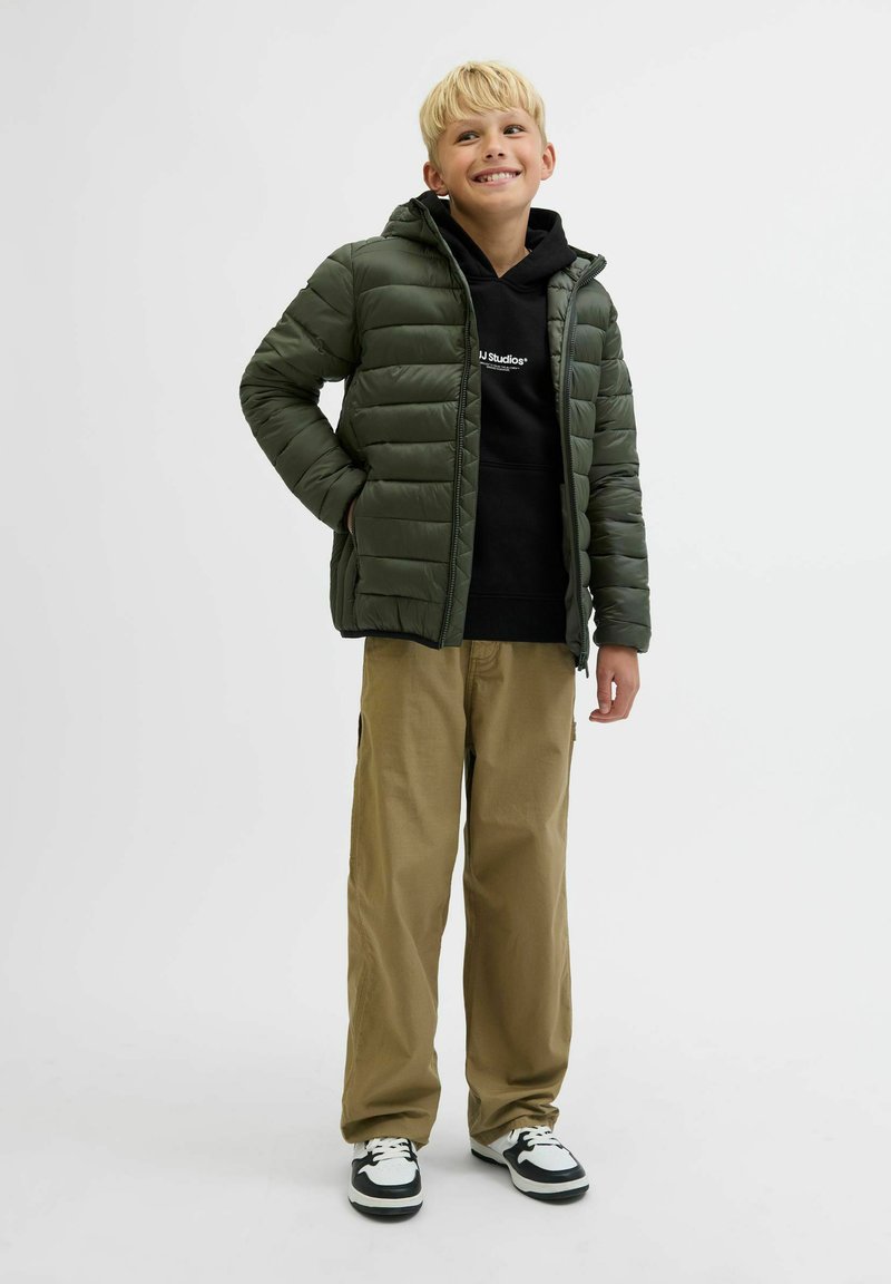 Grön vadderad jacka med huva, svart hoodie under, khakifärgade byxor och svart-vita sneakers. Har en kviltad design och fickor.