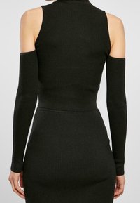 Kvinna i en figurnära svart ribbad ärmlös klänning med avtagbara långa svarta ärmar, som visar bar överkropp och står mot en neutral bakgrund.