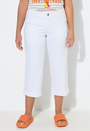 Person trägt weiße, gekürzte Hosen, ein gestreiftes Hemd mit Hummer-Motiv und leuchtend orangefarbene Sandalen und steht vor einem einfarbigen Hintergrund.