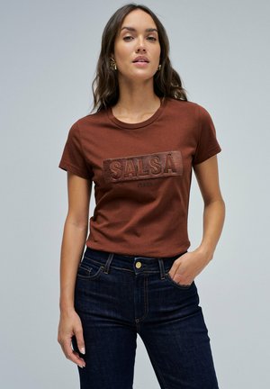 Camiseta de algodón marrón con un logo texturizado "SALSA", de mangas cortas y cuello redondo, combinada con jeans de mezclilla azul oscuro.