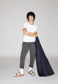 Witte T-shirt met korte mouwen, grijze trainingsbroek, bruine slippers met witte sokken en een donkerblauwe jas over één arm gedrapeerd.