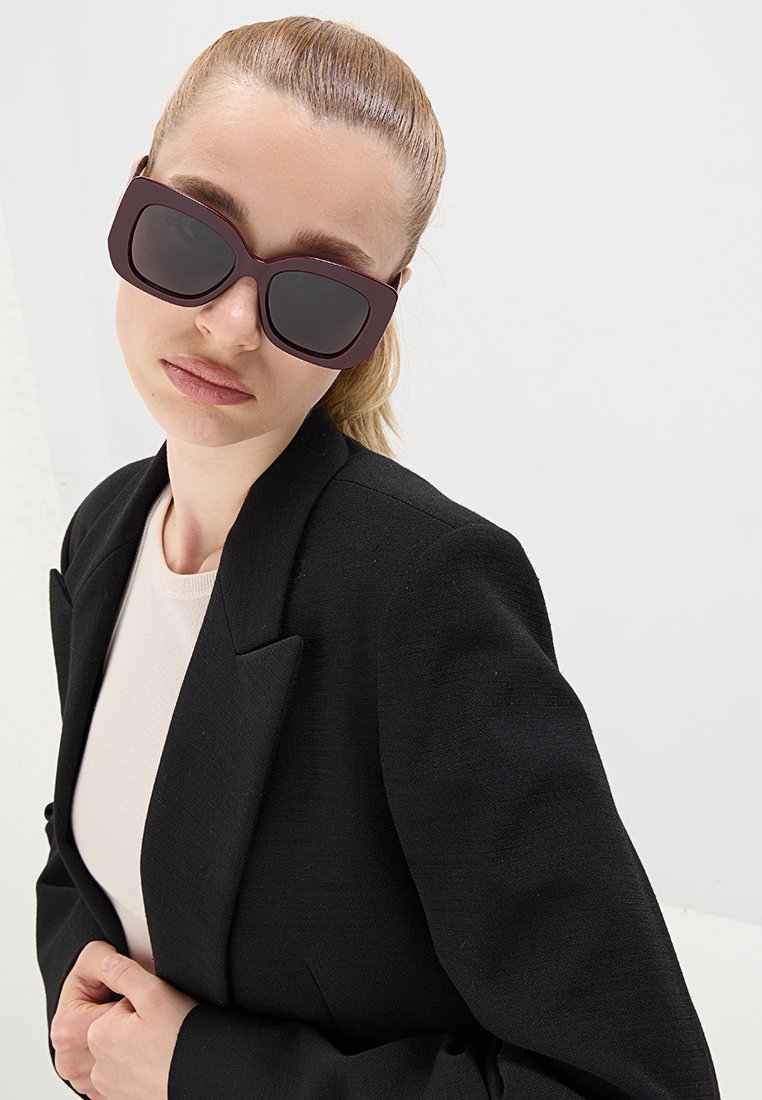 Donna con capelli biondi raccolti in un ponytail, che indossa occhiali da sole oversize neri, un blazer nero e un top beige, posando contro uno sfondo bianco semplice.