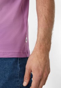 T-shirt in cotone viola con maniche corte, etichetta sul lato e una superficie liscia; indossato con jeans denim scuri, mostrando parzialmente una mano.