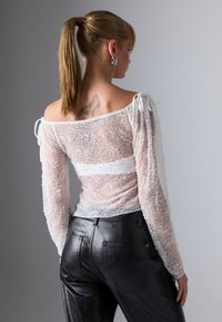 Valge pitsist pikkade varrukatega särk, off-shoulder disainiga, millel on lipsud varrukatel ja läbipaistev tekstuur, kombineeritud mustade nahkpükstega.