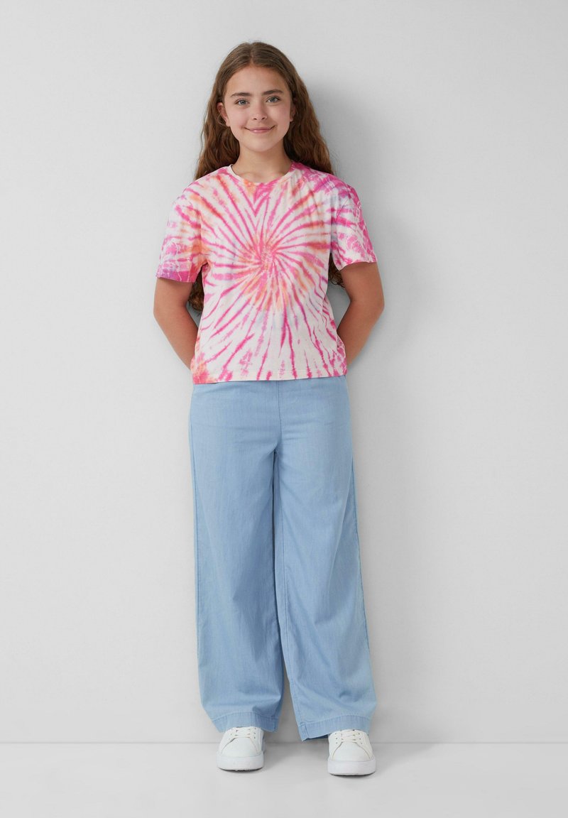 Koszulka tie-dye w różowym i pomarańczowym kolorze ze spiralnym wzorem, w połączeniu z jasnoniebieskimi szerokimi spodniami i białymi trampkami.