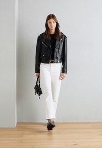Giubbotto biker corto in pelle nera con zip argentate, top nero, pantaloni bianchi a gamba dritta, borsa nera e scarpe grigie chunky su pavimento in legno.