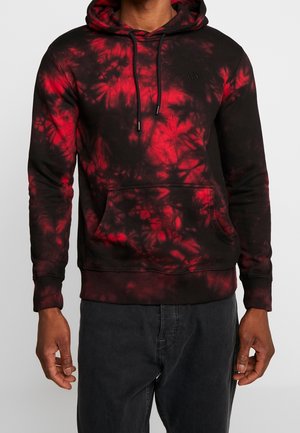 Mann trägt einen rot-schwarzen Batik-Hoodie mit Fronttasche und schwarzen Kordeln, kombiniert mit dunkler Hose vor einfachem Hintergrund.
