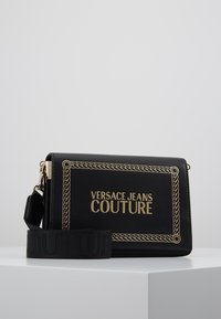 Borsa a tracolla rettangolare nera di Versace Jeans Couture con bordi in catena dorata e logo, dotata di una tracolla in tessuto larga e removibile.