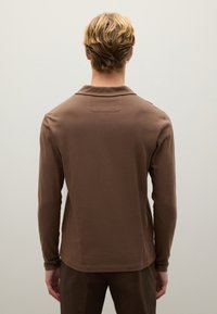 Langärmliges braunes Poloshirt aus weichem Stoff, dezenter Kragen, figurnahe Passform, glatte Textur, minimalistische Nähdetails, getragen mit passenden Hosen.