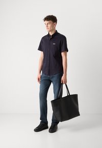 Camicia nera a maniche corte con bottone davanti e logo, abbinata a jeans blu e una grande borsa nera a tracolla. Sneakers nere completano il look.