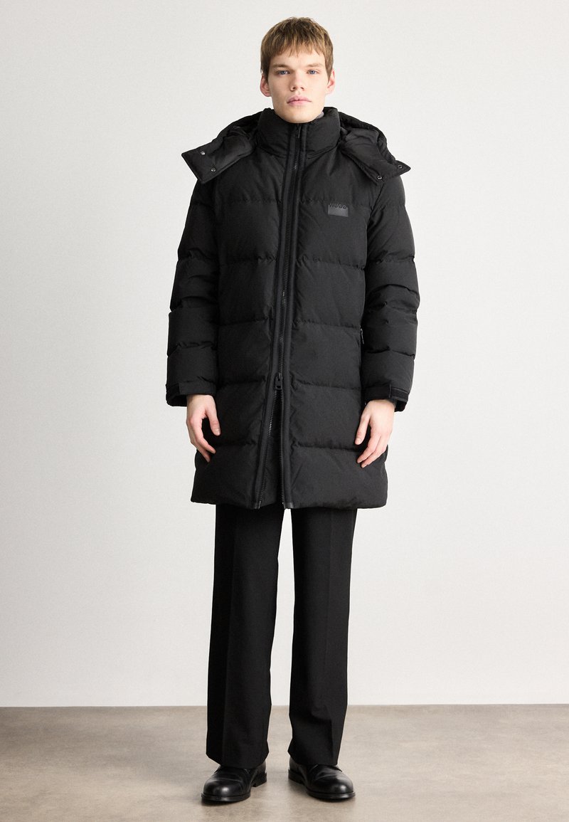 Black puffer coat with a high collar, zip closure, and hood. Features quilted design, side pockets, and a logo patch on the chest.