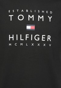 Černé bavlněné tričko s tučným bílým textem: "ZALOŽENO TOMMY HILFIGER MCMLXXXV" s malým logem v červené, bílé a modré barvě.