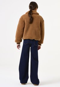 Chaqueta de forro polar marrón con cuello tipo solapa, corte holgado y puños acanalados, combinada con pantalones de mezclilla oscuros de talle alto y pernera acampanada.