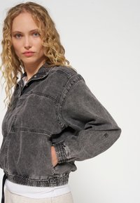 Giacca in denim nero con zip frontale, colletto alto, polsini elastici e vita arricciata. Presenta dettagli cuciti e tasche laterali.