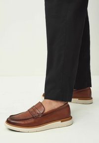 Mocassins en cuir marron avec un design de mocassin, dotés d'une boucle décorative, d'une semelle en caoutchouc beige et de détails de couture subtils. Associés à un pantalon noir.