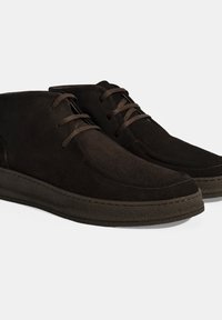 Bruine suède enkelboots met een soepele textuur, ronde neuzen en bruine veters. De rubberen zool heeft een gestikte accent langs de rand.