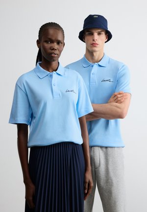 Doi modele poartă tricouri polo bleu Lacoste; femeia își asortează tricoul cu o fustă plisată bleumarin, iar bărbatul poartă pantaloni gri și o pălărie bucket bleumarin.